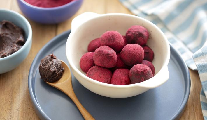 Fotografia em tons de roxo, em uma bancada de madeira marrom. Ao centro, uma tábua redonda azul e no meio um prato branco contendo as trufas. Ao lado, uma colher de madeira e ao fundo, há pó de beterraba em um prato.