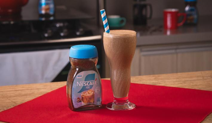 Fotografia de um copo de vidro grande com shake de café, aveia e banana, ao lado de uma embalagem de nescafé gelado, sobre um pano vermelho.
