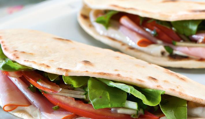 Fotografia em tons de rosa em uma mesa de madeira com um pano rosa florido, uma tábua de madeira branca ao centro com a piadina, que é uma massa estilo crepe francês, recheada de tomate, rúcula e peito de peru.