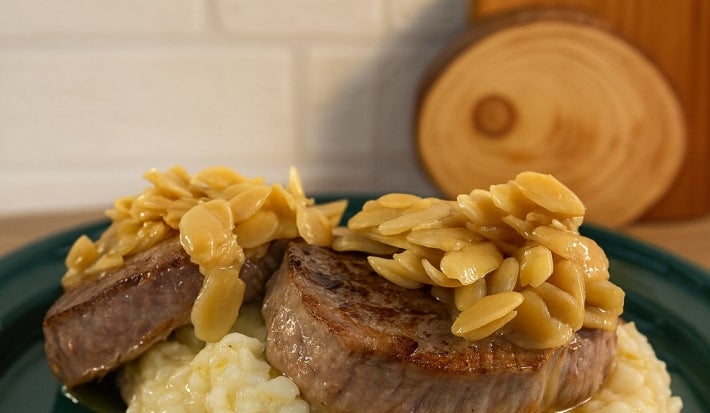 imagem mostra um prato de risoto cremoso em tons beges e por cima 2 medalhões de filé mignon com amendoas laminadas e um molho amanteigado por cima.