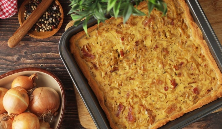Fotografia de uma torta recém-assado em uma cozinha rústica, com cebolas caramelizadas e ervas, em uma bandeja dourada. Ao lado, cebolas inteiras e pimenta em grãos, criando um ambiente acolhedor e caseiro.