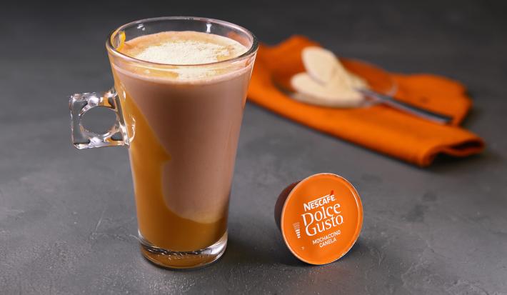 Fotografia em tons de laranja em uma bancada de madeira cinza escura, um paninho laranja e uma xícara de vidro alta com o café de mochaccino canela com doce de leite e achocolatado Galak.
