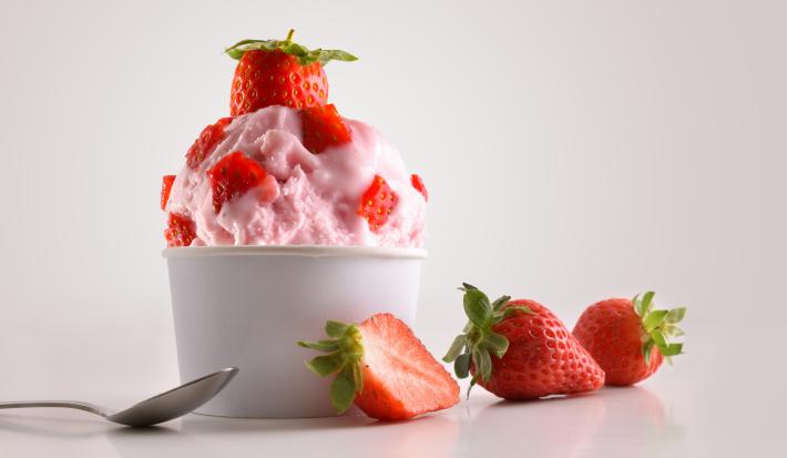 Fotografia de frozen iogurte de morango em um potinho de papel com pedaços de morango, e em cima, um morango inteiro. Ao lado, dois morangos inteiros e um morango cortado pela metade, no outro lado, uma colher de sobremesa, sobre um fundo branco.