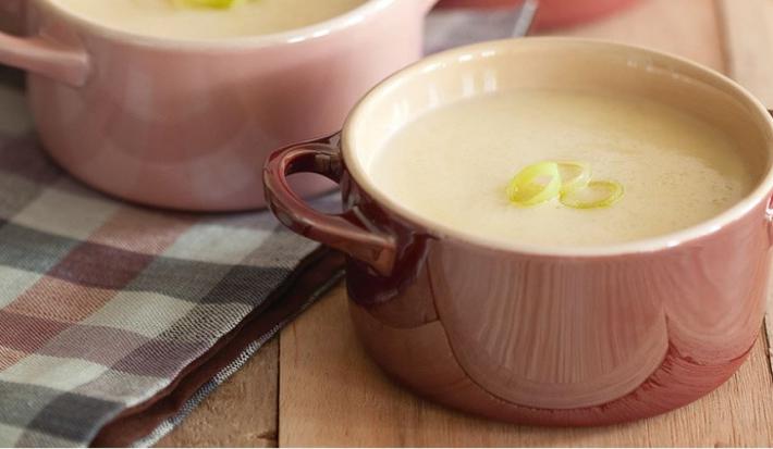 Fotografia em tons de rosa em uma bancada de madeira com uma toalha xadrez preta e rosa, duas mini panelinhas de cerâmica rosa com a sopa de alho-poró dentro delas.