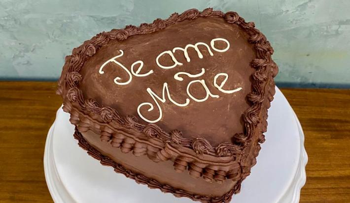 Fotografia em tons de castanho com um prato branco ao centro. Em cima do prato existe um bolo alto em formato de coração com cobertura de chocolate. Em cores brancas existe um escrito "Te amo Mãe"