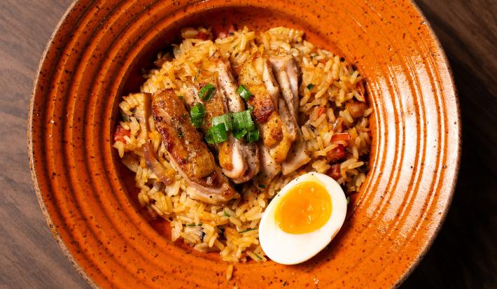 Fotografia vista de cima de uma receita de um arroz frito com uma sobrecoxa de frango em fatias, e pra decorar, cebolinha e uma metade de um ovo com gema mole por cima. A receita está em um prato fundo laranja sobre uma mesa de madeira.