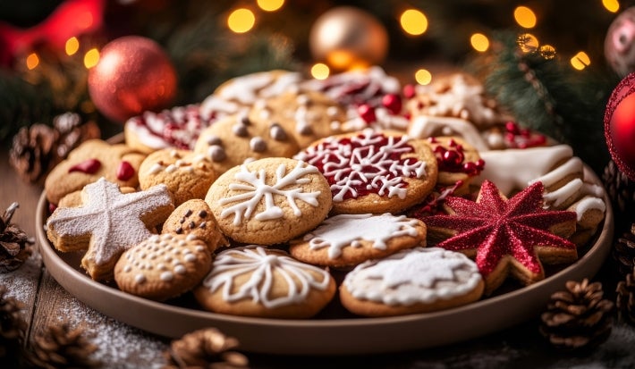 Em cima de uma mesa decorado com enfeites de natal, prato com biscoitos em formato de estrelas e redondos, decorados com glacê branco e vermelho.