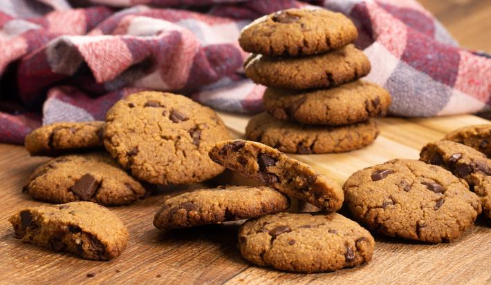 fotografia em tons de marrom, vinho e roxo de uma bancada marrom vista de frente, contém diversos biscoitos de chocolate espalhados pela mesa, e ao fundo  um pano xadrez em tons de vinho e roxo.