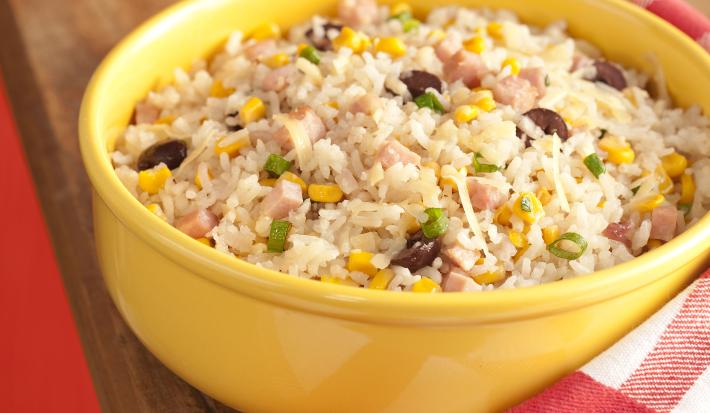fotografia em tons de vermelho, marrom e amarelo de uma bancada marrom vista de frente, acima da bancada contém um recipiente redondo amarelo com arroz peito de peru e milho. Ao lado um pano xadrez em tons de vermelho e branco.