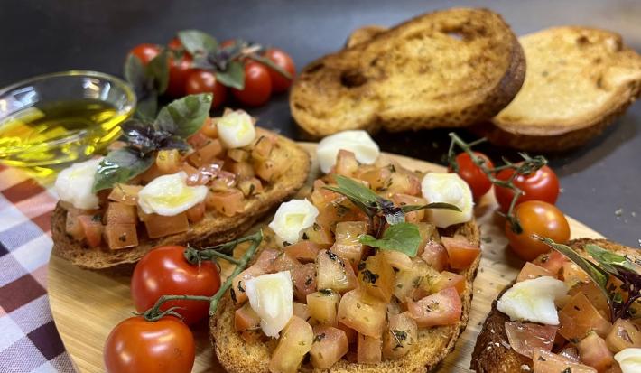 Foto da receita de brusqueta fácil em tons de bege servida em 3 porções dispersas em uma tábua de madeira oval sobre uma mesa quadriculada de cantina. Ao fundo, algumas fatias de pão italiano, tomates e em um recipiente com azeite para decorar