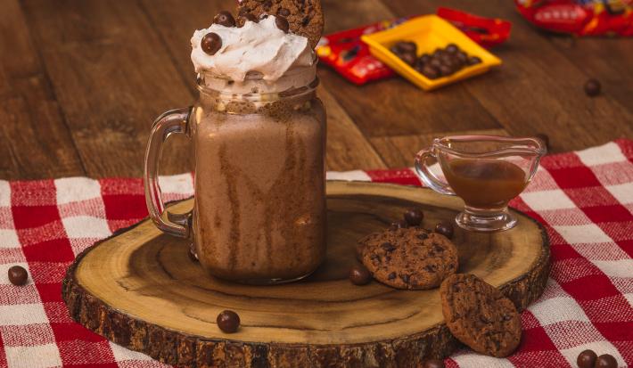 Foto de uma bancada de madeira. Sobre ela há uma toalha quadriculada vermelho e branco, uma tábua de madeira com um copo com o Milkshake de Nescau decorado com um canudo vermelho. Ao lado há um potinho com calda, 2 cookies e algunmas bolinhas de chocolate