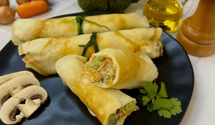 Foto em tons de amarelo da receita de panqueca de legumes cremosa servida em 4 porções sobre um prato largo preto com cogumelos ao lado. Ao fundo, há cenouras e cebolas cortadas, além de um brócolis, um vidro de azeite e um moedor de pimenta do reino