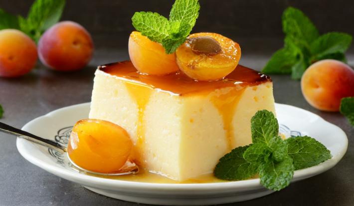 Fotografia de um flan quadrado de pêssego com Leite MOÇA, NESTLÉ Creme de Leite e calda de hortelã sobre um miniprato de vidro. Ao fundo de alguns pêssegos e folhas de hortelã.