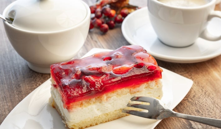 Fotografia de um pedaço de torta de morango em tons claros sobre um praot retangular pequeno branco, apoiado em uma mesa de madeira.