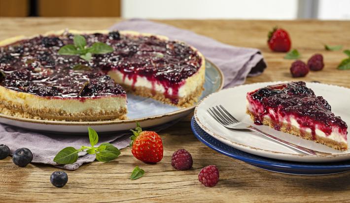 Fotografia em tons de vermelho em uma bancada de madeira com um prato raso grande com a cheesecake de iogurte com frutas vermelhas e um prato raso pequeno com uma fatia da cheesecake. Morangos e uvas espalhadas pela bancada.