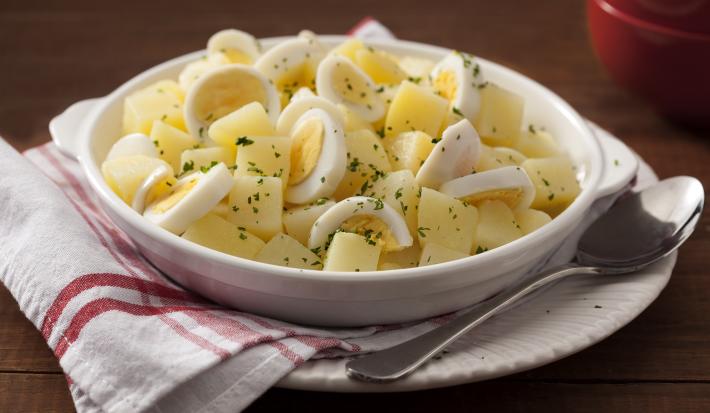 Fotografia em tons escuros e branco de uma bancada de madeira marrom, sobre ela um prato caso branco com um pano quadriculado branco e vermelho, uma colher de inox de uma tigela branca com as batatas e os ovos. Ao fundo, um potinho vermelho.
