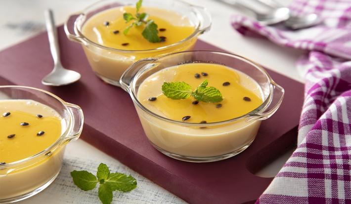 Fotografia em tons de amarelo, em uma bancada de madeira de cor branca. Ao centro, uma tábua de madeira roxa contendo 2 taças de mousse com uma colher ao lado. Ao fundo, há um pano quadriculado roxo.