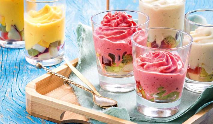 Fotografia em tons de azul, rosa e amarelo em uma mesa de madeira azul com uma bandeja de madeira e copos de vidro com os gelados de fruta, morango e banana. Ao fundo, dois copos de vidro com os gelados de fruta de manga.