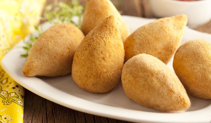 Fotografia em tons de amarelo em uma bancada de madeira, um pano amarelo, uma tábua de madeira, um prato branco oval com várias coxinhas de frango em cima dele. Ao fundo, um potinho com ketchup.