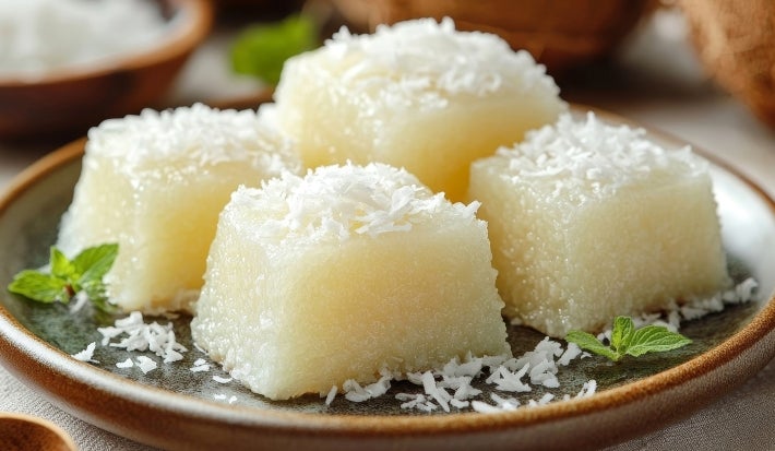 Fotografia de um prato rústico com pedaços de sobremesa de tapioca, cobertos com coco ralado e decorados com folhas de hortelã, em um ambiente tropical, com um fundo que sugere mais ingredientes de coco.