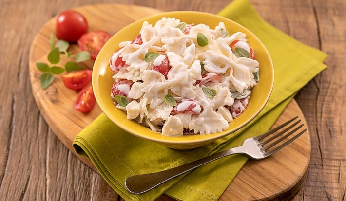 Foto da receita de saladinha de macarrão no copinho servida em um copo largo amarelo sobre um paninho verde com um garfo prateado ao lado, há também, em cima da tábua de madeira, tomates cerejas cortados ao meio