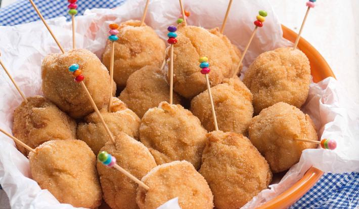 Fotografia em tons de azul em uma bancada de madeira clara com um pano azul quadriculado e em cima um prato redondo fundo laranja com coxinhas cremosas de frango dentro dele e palitinhos coloridos espetados nelas.
