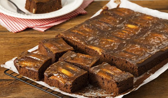 fotografia em tons de marrom e branco de uma bancada marrom vista de frente. Contém uma grade com um papel branco por cima, que contém um bolo de chocolate com pedaços de maçãs e cortado em quadrados