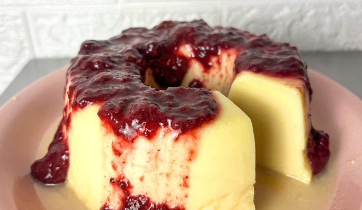 Fotografia em tons claros com um pudim de NINHO coberto com geleia de frutas vermelhas ao centro. O pudim possui cor clara e a geleia de frutas vermelhas escorre delicadamente sobre o pudim.