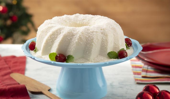 Foto da receita de Sobremesa Gelada de Coco. Observa-se uma boleira azul com a sobremesa em cima, polvilhada de coco e enfeitada com cerejas na base. Decorações de Natal enfeitam a foto, como a árvore no fundo desfocada e bolas natalinas.