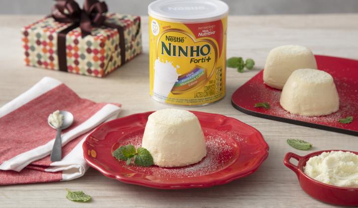 Fotografia em tons de vermelho e amarelo de uma bancada de madeira, ao centro um prato vermelho com uma porção do Brigadeirão de Caneca, uma lata de NINHO Forti+ e ao fundo um pacote de presente e mais brigadeirões.