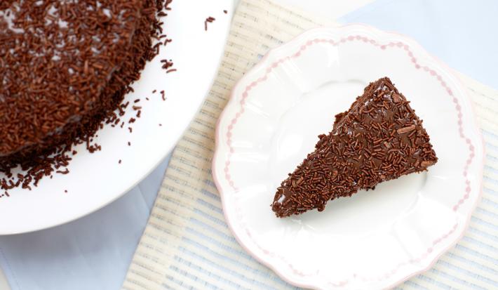 Imagem vista de cima de um prato branco com detalhes rosa e uma fatia de bolo de chocolate com brigadeiro e chocolate granulado.