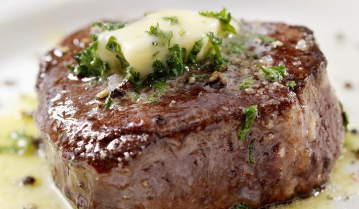 Fotografia de um bife mais alto feito, em um prato branco, com um azeite de ervas e pimenta sobre a carne e escorrendo no prato. Em cima da carne, manteiga e salsinha.
