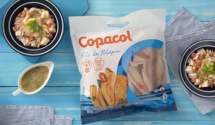 Foto da receita de Ceviche Refrescante com Filé de Tilápia Copacol, vista de cima, servido em uma tigela azul, ao lado de uma molheira branca, sobre uma bancada azul com tecidos e com um pacote do produto Copacol