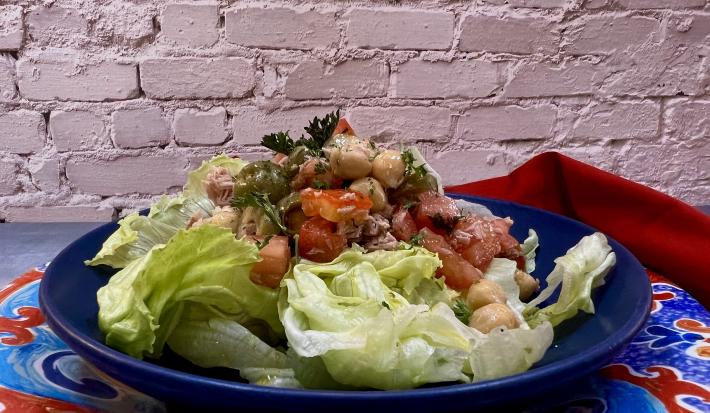 Foto da receita de Salada de Grão de Bico. Observa-se um prato azul redondo fundo com a salada com tomate, grão de bico, atum e alface americana dentro. Ao fundo, uma parede de tijolo branco.