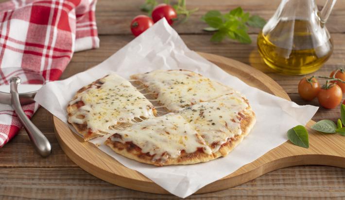 Fotografia em tons de amarelo e bege de uma bancada de madeira, ao centro uma tabua de madeira com um pedaço de papel manteiga e um disco de pizza com molho e queijo derretendo. Ao fundo um pano xadrez vermelho e branco e um vidro com azeite.