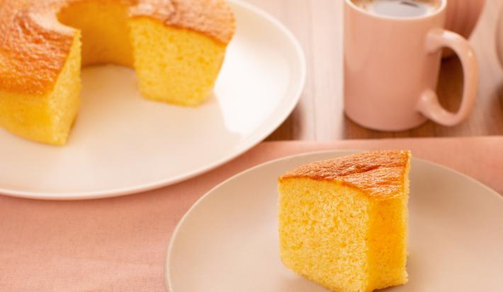 Fotografia de um prato raso branco grande com um bolo quase inteiro de fubá, na frente tem outro prato, menor e de tom mais escuro, com uma fatia do bolo de fubá. Um pouco mais ao fundo tem uma xícara rosa com café.