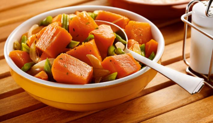 Fotografia em tons de laranja em uma mesa de madeira com um potinho redondo laranja e o refogado de abóbora e vagem dentro. Ao fundo, uma travessa com arroz e ao lado um saleiro.