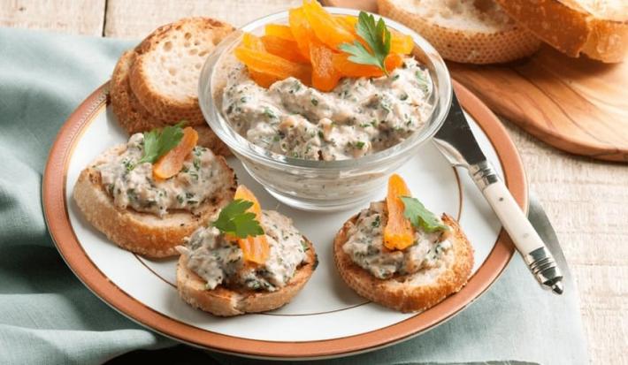 Fotografia em tons de laranja em uma bancada de madeira com um pano cinza, um prato branco com detalhes laranja vários canapés com patê de atum e damasco. Ao centro, um potinho de vidro com o patê de atum e damasco.