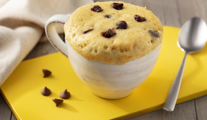 Fotografia em tons de marrom. branco e amarelo de uma bancada de madeira com uma tábua amarela, sobre ela uma xícara branca com bolo de caneca e uma colher. Ao fundo um paninho branco.
