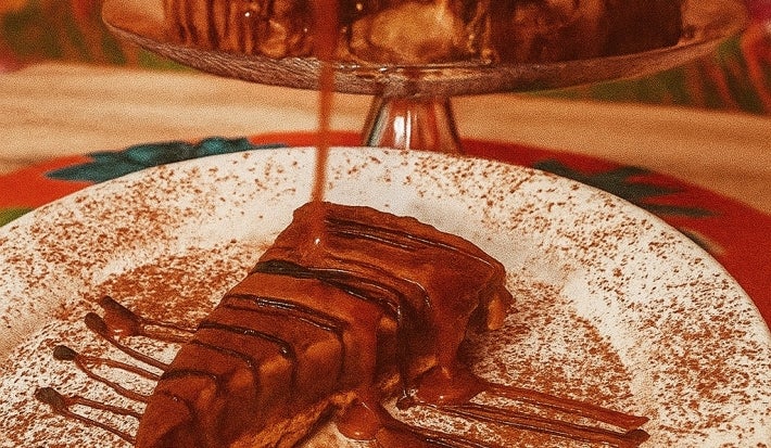 Fotografia de uma fatia de torta de ninho e chocolate em um prato branco raso salpicado de cacau em pó. Por cima da fatia tem calda.