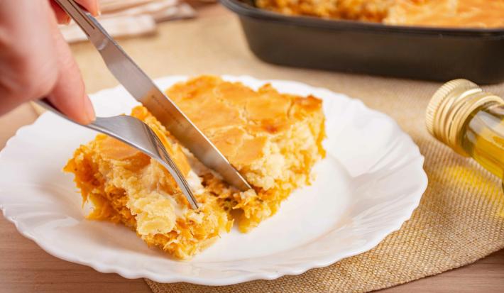 Fotografia de uma fatia de trota de frango em um prato raso de vidro branco. Tem uma mão masculina cortando um pedaço com garfo e faca. No fundo tem a uma forma preta com a torta assada.