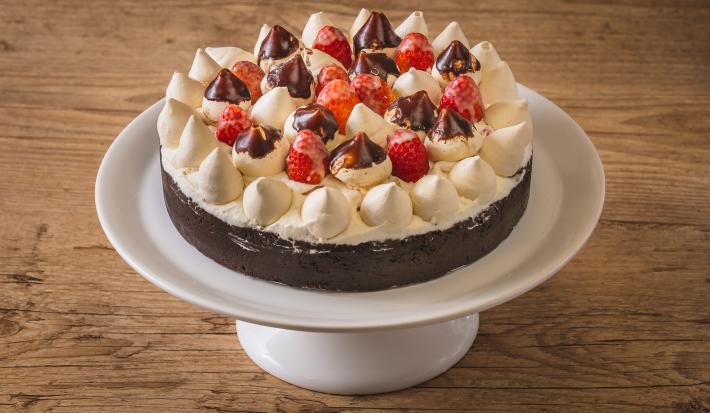 Foto aproximada de uma torta num prato branco alto. A torta tem uma base de biscoito escura e está decorada com chantilly, suspiros pequenos e morango, alguns banhados em chocolate. Tudo está numa bancada de madeira.