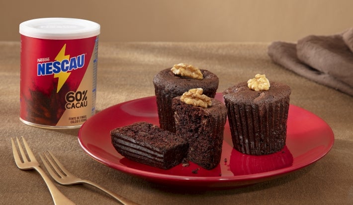 Fotografia de três muffins de nescau sobre um prato vermelho, raso, ao lado de dois garfos dourados e uma lata de NESCAU Extra Cacau. Um dos muffins está cortado na metade para mostrar a maciez da massa.