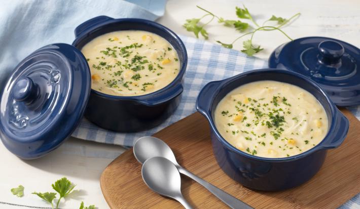 Fotografia em tons de branco, azul e marrom de uma bancada de madeira branca com um tábua de madeira, sobre ela um recipiente redondo azul com a sopa e duas colheres. Ao lado um paninho xadrez branco e azul com um recipiente azul com a sopa.