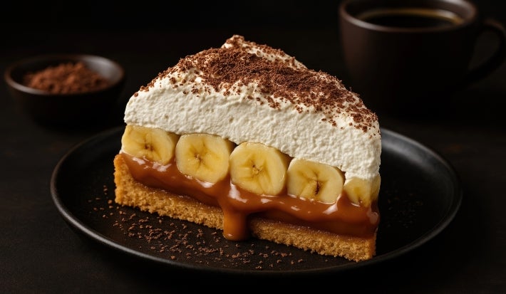 Em cima de uma mesa, prato com fatia de banoffee com massa de biscoitos crocante, doce de leite, bananas em rodelas, decorado com chantilly cremoso e cacau em pó.