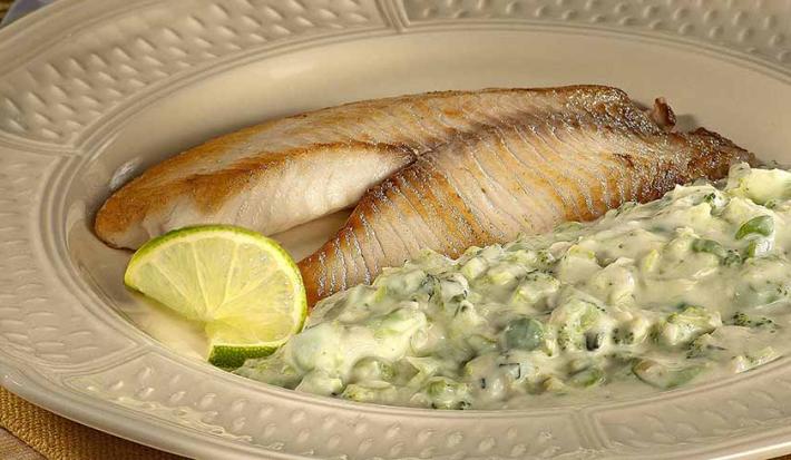 Fotografia em tons de verde em uma mesa de madeira, com um jogo americano amarelo, um prato redondo bege com o filé de peixe e o purê de brócolis dentro dele.