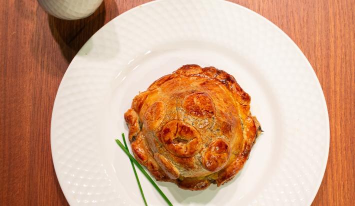 Fotografia de uma receita de pithivier paraense com duxelles de ora-pro-nóbis sobre um prato raso branco, o qual está sobre uma bancada de madeira. Ao lado do prato tem um recipiente fundo e pequeno com molho.