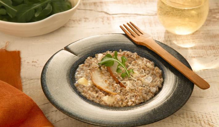 Foto da receita de risoto de pera com brie servida em um prato fundo azul com um garfo de madeira na borda e um copo de vidro com vinho branco ao fundo