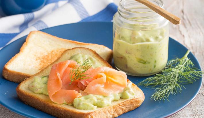 fotografia em tons de cinza, azul e verde tirada de uma bancada cinza, com um prato azul e redondo que contém 2 pães de forma e um deles contém salmão e guacamole. Ao lado esquerdo do prato contém um potinho com a guacamole dentro.