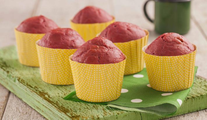 Fotografia em tons de verde em uma bancada de madeira clara, uma tábua de madeira verde, um paninho verde com bolinhas brancas e forminhas amarelas de muffin com o bolinho de beterraba dentro delas. Ao fundo, uma xícara verde